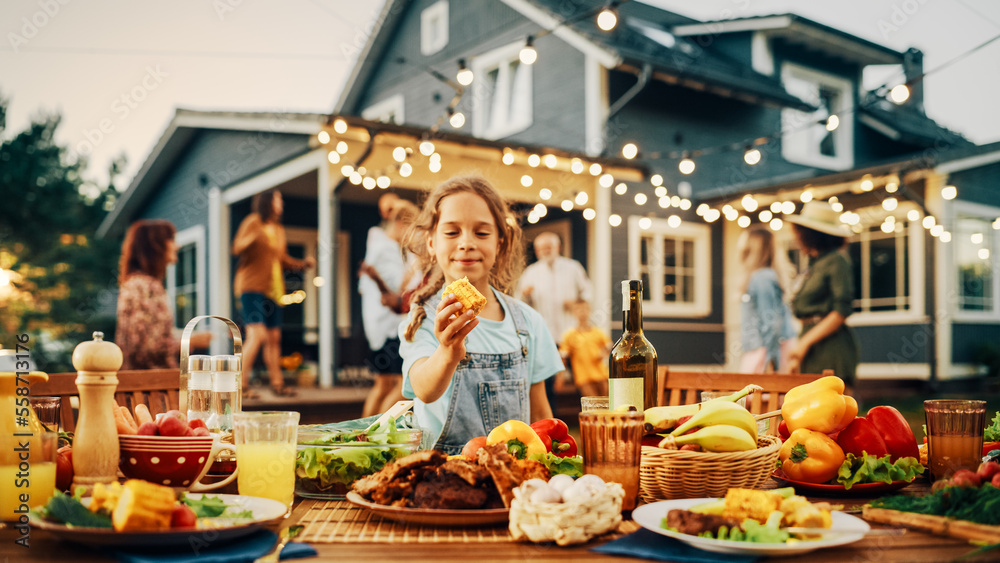 Outdoors Dinner Table with Delicious Barbecue Meat and Fresh Vegetables and Salads. Little Girl Eating a Grilled Corn. Happy People Dancing and Having Fun in the Background.