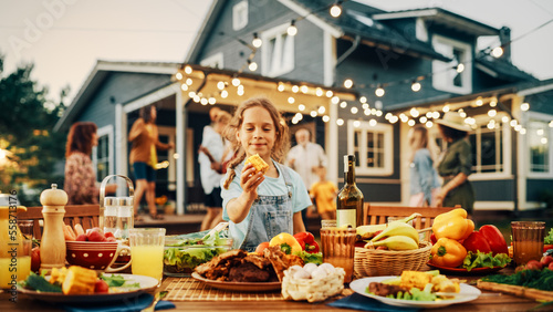 Outdoors Dinner Table with Delicious Barbecue Meat and Fresh Vegetables and Salads. Little Girl Eating a Grilled Corn. Happy People Dancing and Having Fun in the Background.