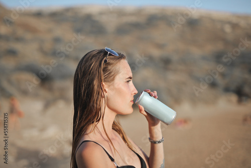 young woman drinks water from a can at the beach