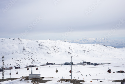 Snow-capped mountain ski slopes with ski lifts against cloudy sky and village in the valley. Winter vacation. Extreme sport and travel content. © DiandraNina