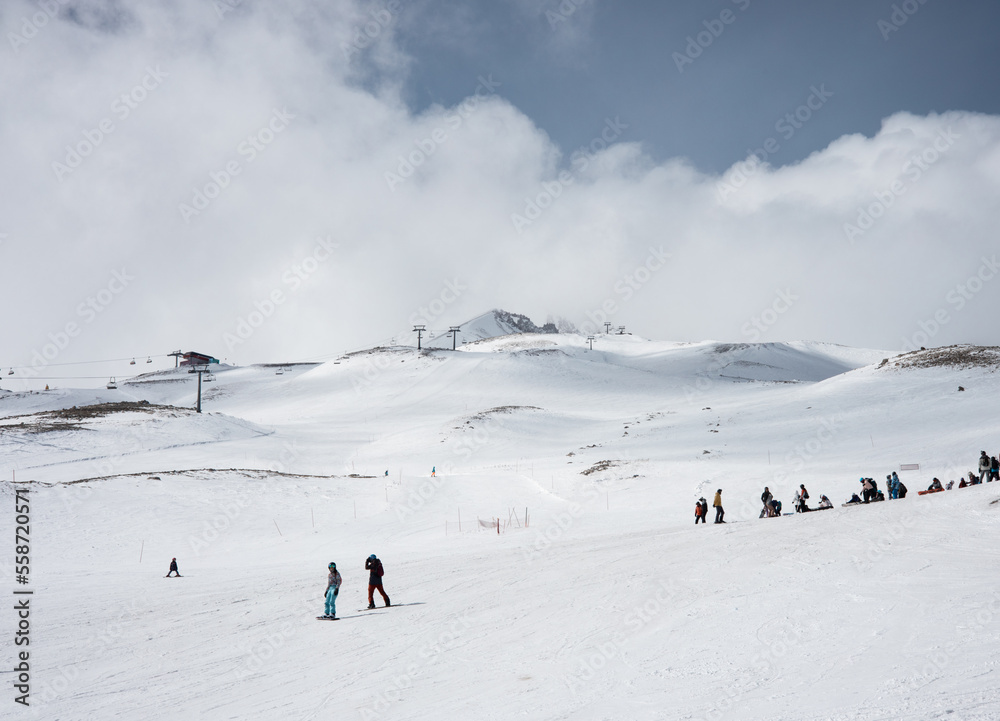 Skiers go downhill skiing on snow-covered mountain ski slope against the backdrop of a cloudy sky and ski lifts. Winter. Extreme sport and travel content  