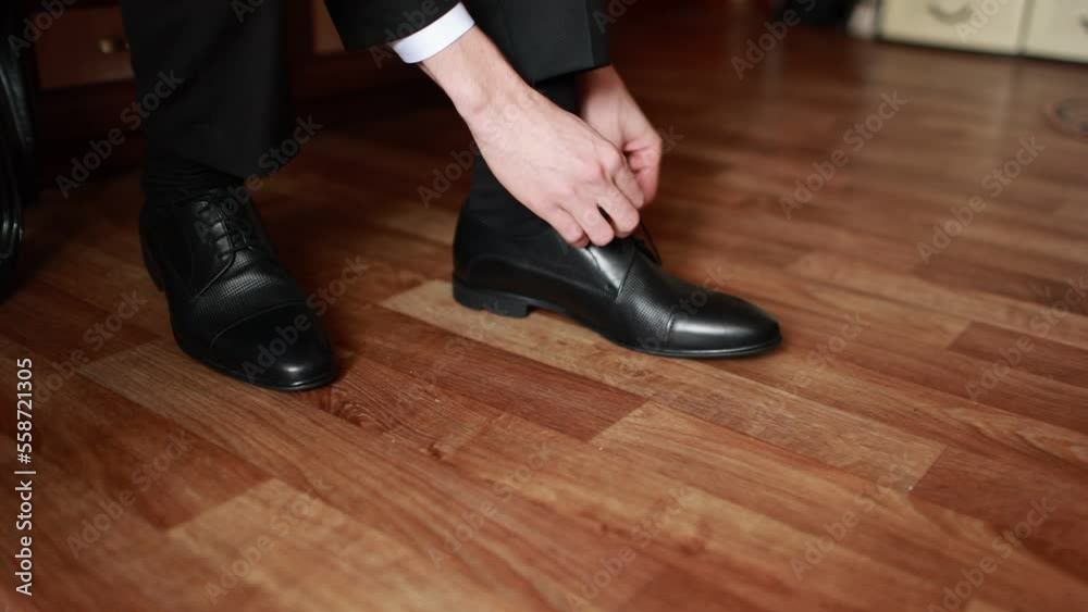 A man in a suit ties up shoelaces on black leather shoes on a wooden parquet background business style. preparation for a wedding or business meeting. means for care of leather shoes A man in a suit ties up shoelaces on black leather shoes on a wooden parquet background business style. preparation for a wedding or business meeting. means for care of leather shoes