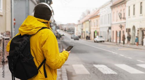 Tableau sur toile Man in travel yellow jacket and headphones standing on roadside in city holding smart phone in his hand and waiting taxi