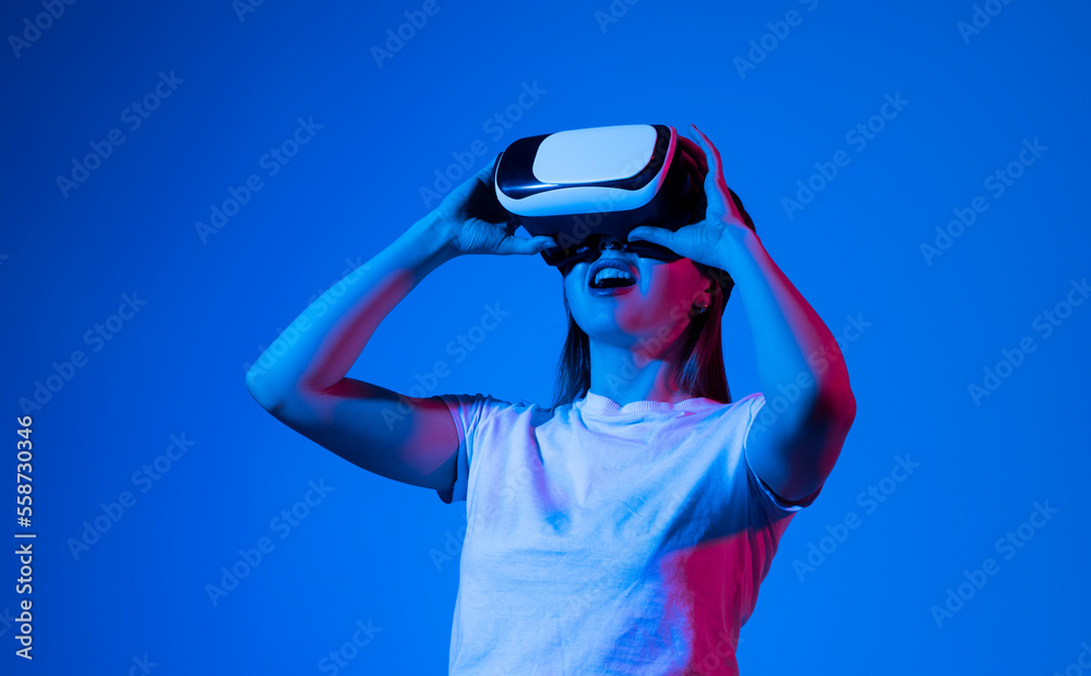 Metaverse concept of young brunette woman in white t-shirt wearing VR headset and playing video games and interacting with virtual reality in neon light.