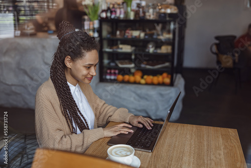Young African woman concentrated on working remotely, typing text on laptop, sitting in cafe against showcase background
