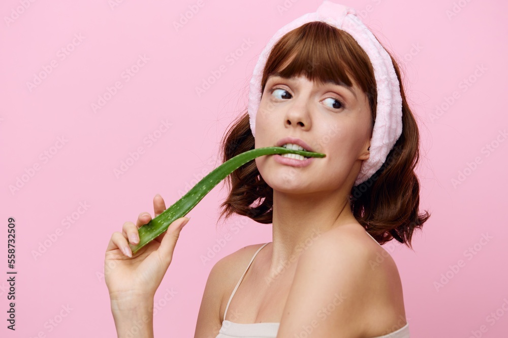 Horizontal studio shot. a cute woman with healthy clean skin and dark beautiful hair stands on a pink background with a cute bandage on her head and bites an aloe leaf. Horizontal studio shot. a cute woman with healthy clean skin and dark beautiful hair stands on a pink background with a cute bandage on her head and bites an aloe leaf.