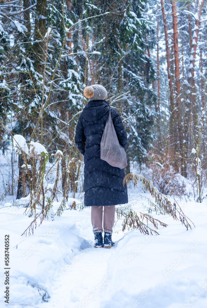 Woman standing her back in winter forest during holiday stroll, walk in woods alone with tote bag, in knitted hat, puffer jacket
