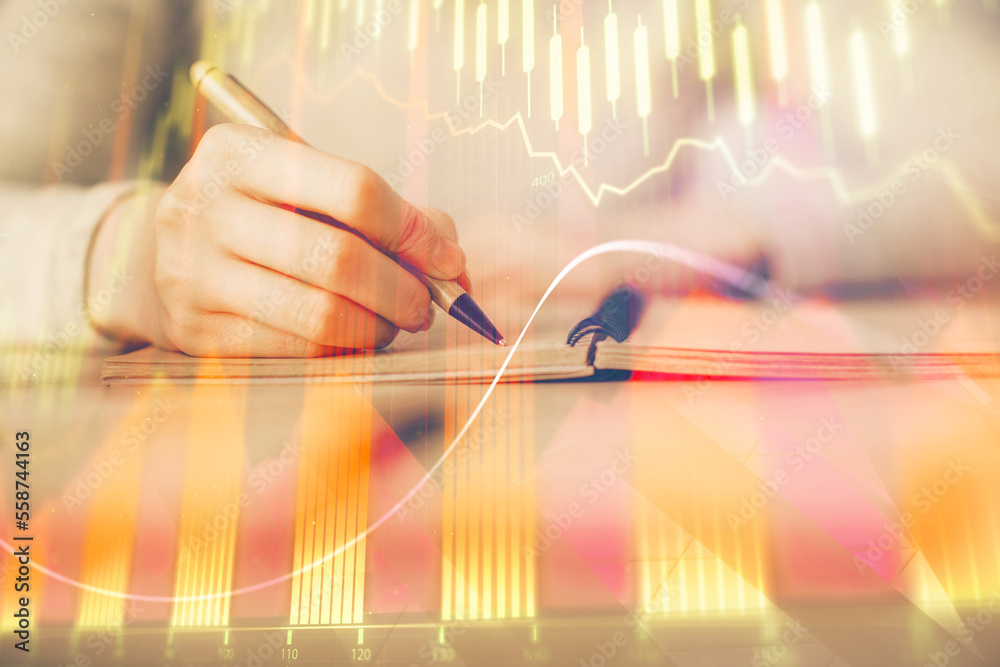 Fototapeta premium Hands writing information about stock market in notepad. Forex chart holograms in front. Concept of research. Multi exposure
