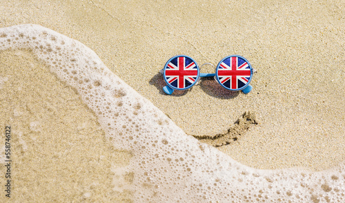 Sunglasses with flag of Great Britain on a sandy beach. Nearby is a sea lightning and a painted smile. Travel and vacation concept for Britons