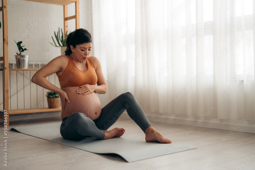 Young pregnant woman suffering from backache and having contractions while sitting on the floor at home.