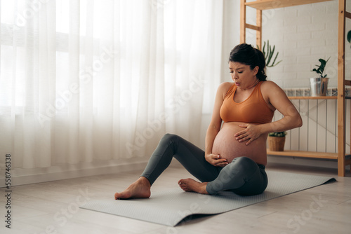Pregnant woman having an unexpected abdominal pain during workout at home. Breathing deeply and holding her belly.