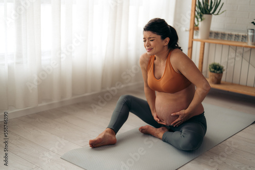 Unhappy Caucasian expecting woman suffering from abdominal pain while sitting on a exercise mat at home.