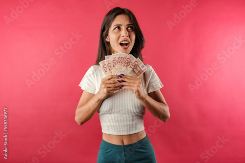 Beautiful brunette woman wearing white ribbed crop isolated over red background holding the banknotes or money looks up and shouts. Lost money or missed a discount.