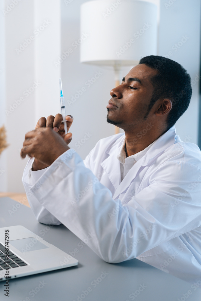 Vertical shot of African-American male doctor in white uniform thoughtful looking to syringe with vaccine in hand sitting at table with laptop in clinic. Concept of vaccination, immunization Vertical shot of African-American male doctor in white uniform thoughtful looking to syringe with vaccine in hand sitting at table with laptop in clinic. Concept of vaccination, immunization