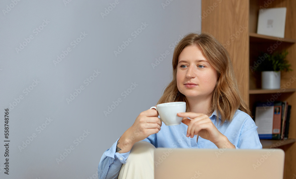 Beautiful young woman working on laptop while sitting at the living room, drinking coffee.
