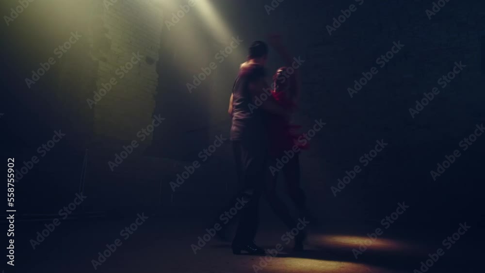 Silhouettes of ballroom dancers pair in darkness of abandoned loft dancing tango under rays of diffused daylight coming from above aside. Passionate synchronous hand movements, rotations and support. Silhouettes of ballroom dancers pair in darkness of abandoned loft dancing tango under rays of diffused daylight coming from above aside. Passionate synchronous hand movements, rotations and support.