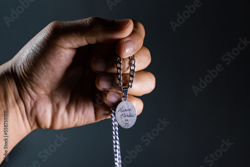 Mano sosteniendo collar con placa que tiene grabada la frase “nunca pierdas la fe” en fondo negro y blanco y negro. photo