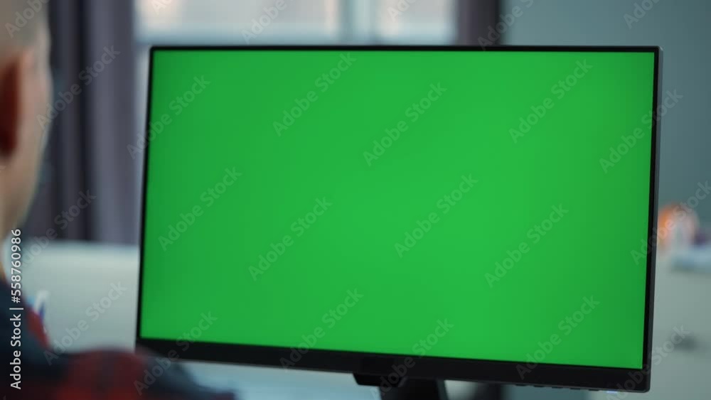 Young Man Working At Computer With Green  Mock Up Screen  Sitting At Desk in Office. Desktop Computer Monitor with Mock Up Green Screen Chroma Key Display Standing on Desk