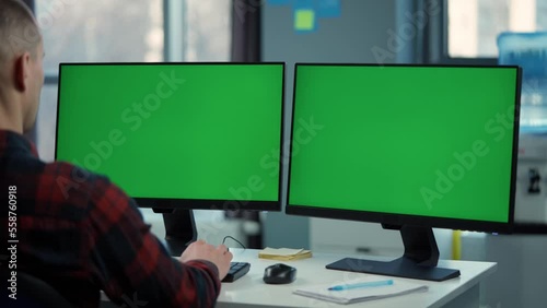 Young Man Working On Computer With Two Green Screen Mock Ups Sitting At Desk In Office