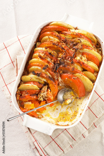 Sliced and layered potato and pumpkin casserole in a white baking form on marble background, topped with roasted onions, feta cheese crumbs and whipped cream