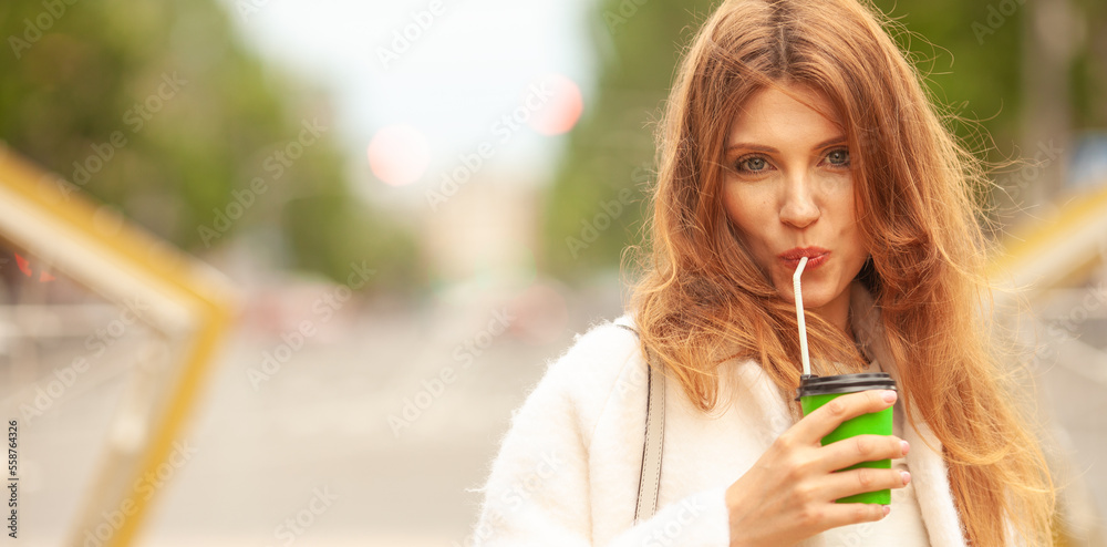 Smiling young woman drinking hot beverage through sipper at street of Mariupol city, Ukraine. Wind blown long hair. Model wearing white coat. Copy-space. Close up outdoor shot