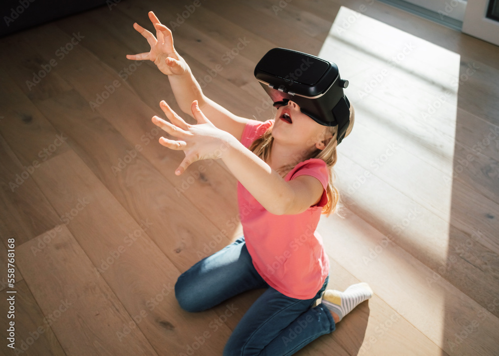 Fototapeta premium Caucasian female kid in a virtual reality headset sitting on the living room floor, looking around excited on the virtual world