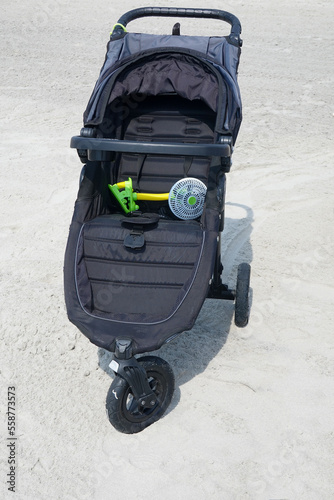 Black three wheel baby stroller with an attachable fan on a white sand background photo