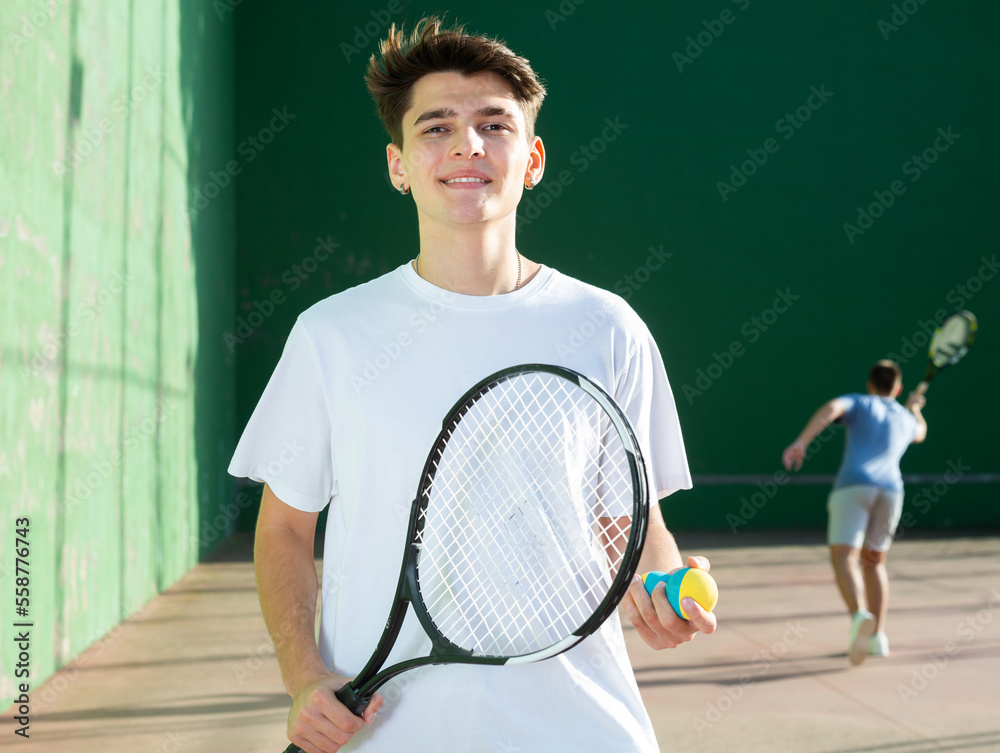 Portrait of positive caucasian boy standing on frontenis court, holding racket and ball