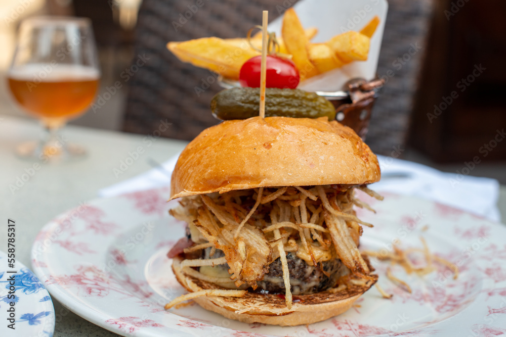 A hamburger with a pickle and tomato attached to the top bun on a blue and white plate with a copper cup filled with French fries. There's a pulled spicy pork sandwich on a plate on the table. 