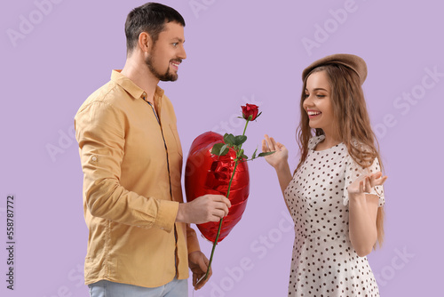 Happy young couple with heart-shaped balloon and flower on lilac background. Valentine's Day celebration