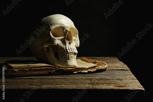 Human skull and old book on wooden table against black background, space for text