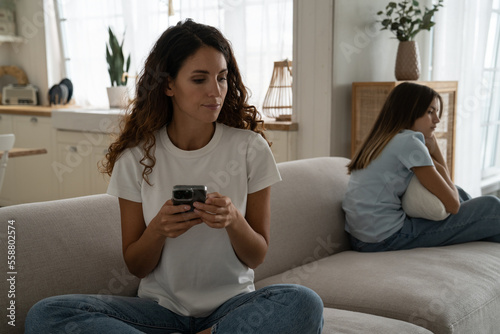 Unhappy sad young caucasian mother and teenage daughter sitting separately on sofa after quarrel. Upset mom holding mobile phone thinking about difficult character of adolescent child ignoring her