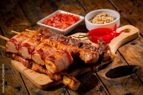 Espetinhos de churrasco em tábua de madeira com acompanhamentos, arroz, vinagrete, farofa e maionese