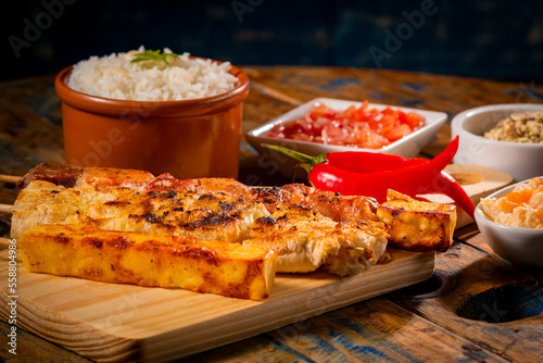 Espetinhos de churrasco em tábua de madeira com acompanhamentos, arroz, vinagrete, farofa e maionese
