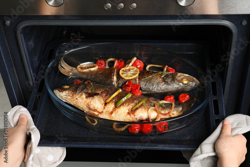 Photography Woman taking out baking tray with sea bass fish and garnish from oven, closeup