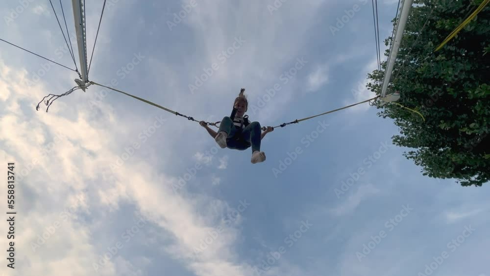 Looking up shot of young little girl having fun jumping and flipping with harness on elastic trampoline. Low angle pov
