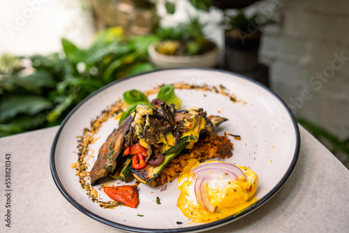 Stack of zucchini  eggplant  and capsicum pan-fried with potato fritters  poached egg  red onion  and cheese sauce on the side. Decorated with chia seeds and avocado and served with a cappuccino.