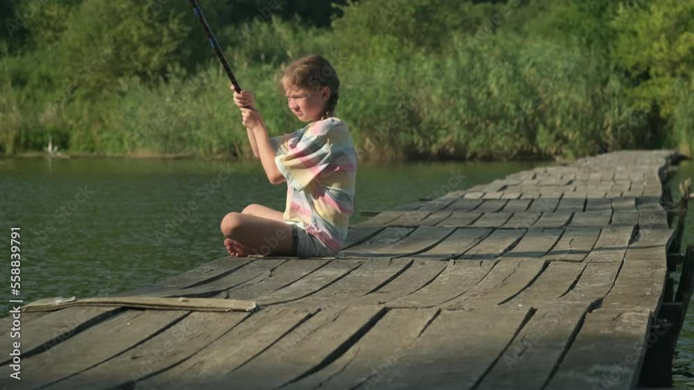 Kid girl is fishing alone on wooden bridge early in the morning. A child fisherman sits with a fishing rod on a river bank and waits for a nibble Kid girl is fishing alone on wooden bridge early in the morning. A child fisherman sits with a fishing rod on a river bank and waits for a nibble