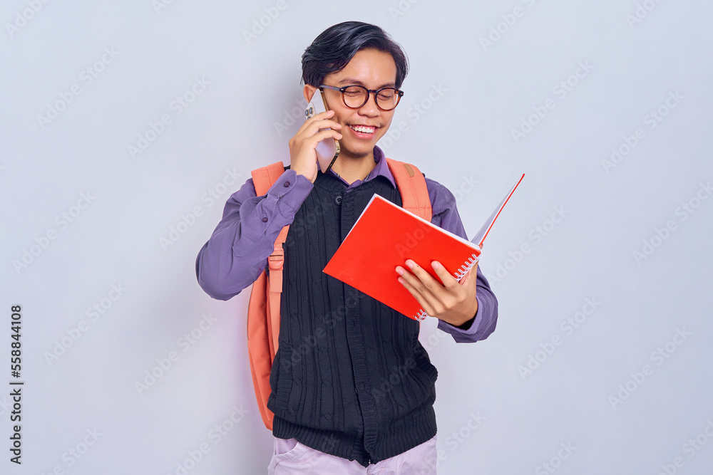 Smiling young Asian student man 20s wearing casual clothes with backpack looking at school books, talking on mobile phone on gray background. Education in high school university college concept Smiling young Asian student man 20s wearing casual clothes with backpack looking at school books, talking on mobile phone on gray background. Education in high school university college concept