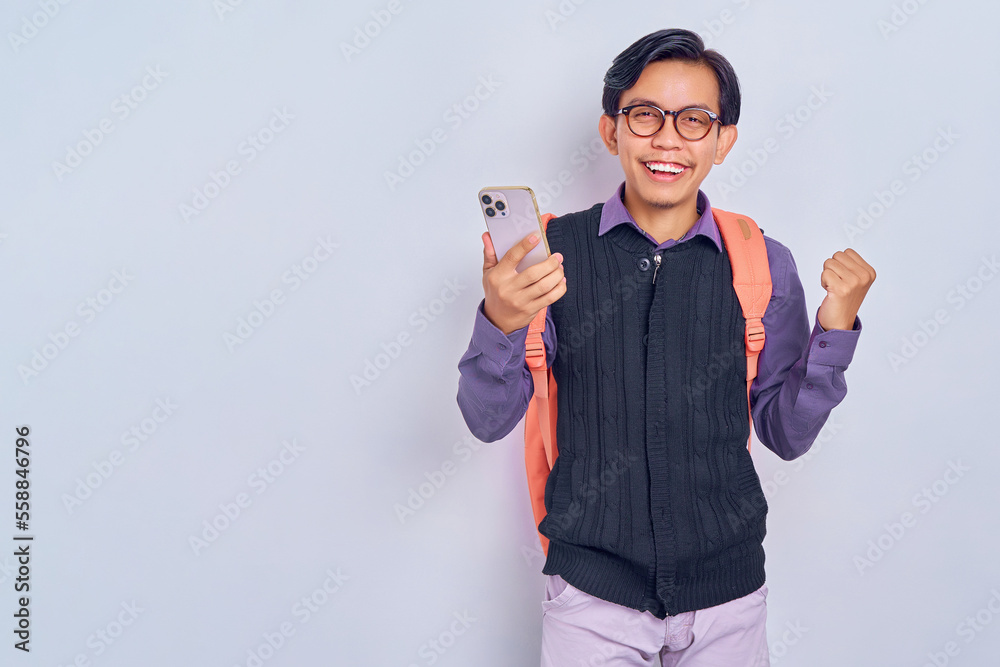Excited young Asian student man in casual clothes with backpack holding smartphone and making victory gesture isolated on gray background. High school university college concept Excited young Asian student man in casual clothes with backpack holding smartphone and making victory gesture isolated on gray background. High school university college concept