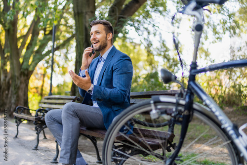 Handsome businessman talking by mobile phone while sitting on bench in park. Bicycle in blur.