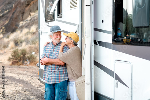 Happy senior couple on a leisure trip standing outside a camper van tenderly embracing. Handsome retired caucasian couple enjoying freedom and alternative life in camper van