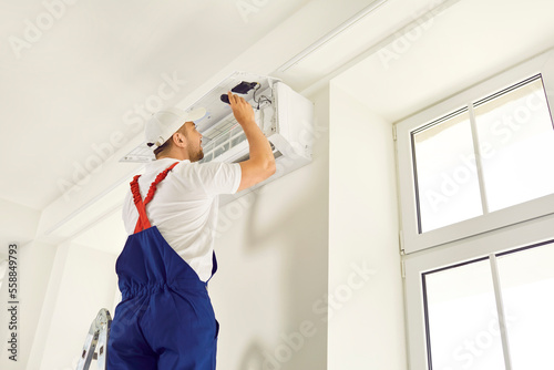 Male technician repairs, cleans or installs air conditioner hanging on wall inside house. Man in work clothes and works on call at client's house. Concept of service and maintenance of air conditioner