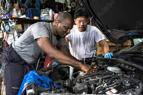 Black mechanic man fixing car in auto repair shop with Asian mechanic assistance, Car Mechanic Concept