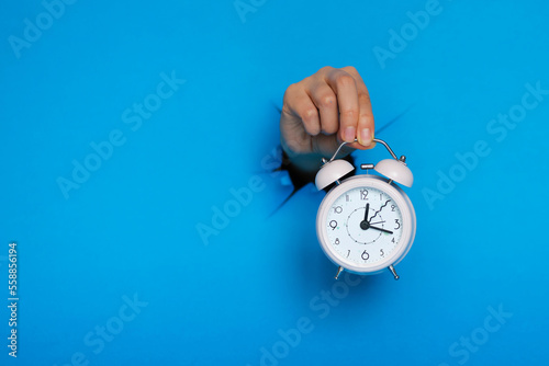 female hand holds pink alarm clock through a paper hole in blue background.