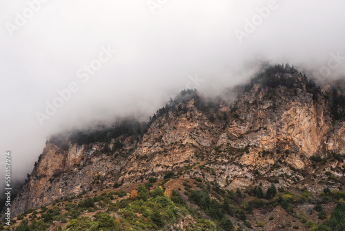 Dark atmospheric landscape with high mountain silhouettes in dense fog in rainy weather. Forest rocky mountain top above hills in thick fog in dramatic overcast. Black rocks in low clouds during rain.