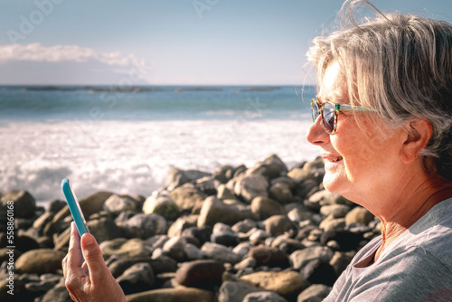 Side view of smiling senior woman using mobile phone while sitting on the beach at sunset - elderly happy lady enjoying free time and retirement at sea #558860168