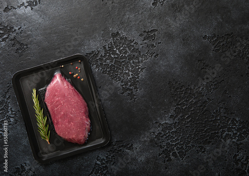 Fotografija Fresh raw beef fillet steak sealed in vacuum tray with pepper and rosemary on black kitchen table background