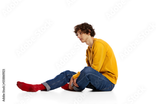 Portrait of young boy sitting on floor in sadness, feeling asocial over white background. Loneliness, depression