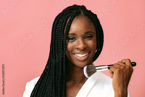 Woman holding make up brush smiling looking at camera on pink background. Beautiful african american young adult